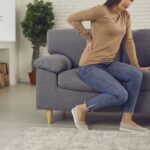 Young woman wants to get up from the sofa but feels a sudden pain in her lower back