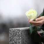 Woman holding white rose near grey granite tombstone outdoors, closeup. Funeral ceremony